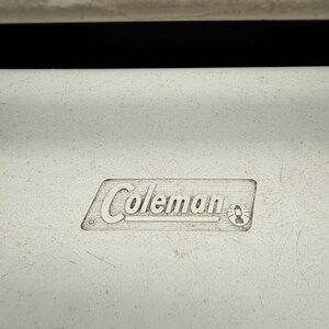 May include: Close-up of a white cooler with the embossed "Coleman" logo. The logo is in a rectangular frame and includes a small lantern icon. The cooler has a textured surface and a black background.