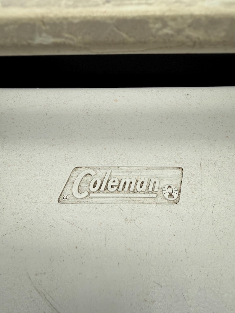 May include: Close-up of a white cooler with the embossed "Coleman" logo. The logo is in a rectangular frame and includes a small lantern icon. The cooler has a textured surface and a black background.
