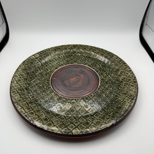 May include: A stack of two decorative plates. The plates have a dark green, textured exterior with a repeating pattern. The center of each plate is a dark brown, circular design. The plates are on a white background.