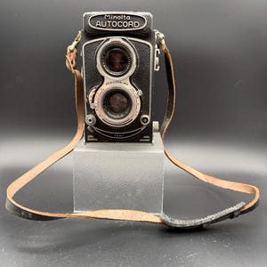 May include: A vintage Minolta Autocord camera with a leather strap. The camera is black with silver accents and features two lenses. The strap is brown. The camera is displayed on a clear stand against a dark background.