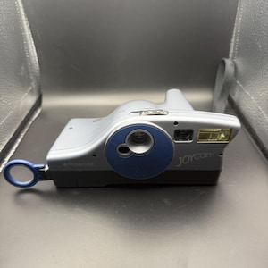 May include: A silver and blue Polaroid Joycam instant camera. The camera has a blue lens ring and a flash on the right side. The word "Polaroid" is printed on the side, and "JOYcam" is printed on the front.