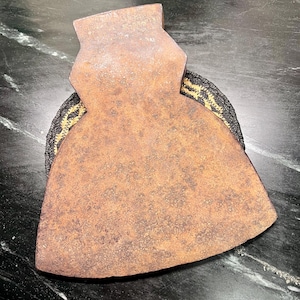 May include: A vintage, rusty axe head with a wide blade and a rectangular top. The axe head is resting on a black and gold woven coaster, set against a dark, marbled surface. The axe head shows signs of age and wear.