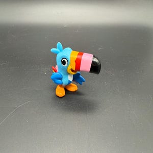 May include: A small, colorful toucan figurine. The bird has a blue body, orange feet, and a large, multi-colored beak with pink, red, yellow, and black accents. The toucan is standing on a dark surface.
