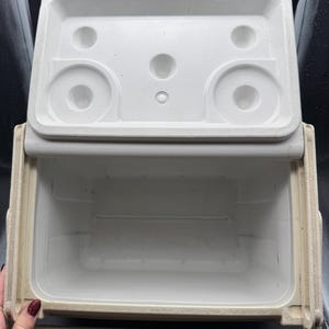 May include: An open, off-white cooler with a matching lid. The lid has circular indentations and a handle. The interior of the cooler is visible, showing a clean, empty space. The cooler is designed for carrying and keeping items cold.