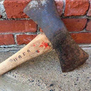 May include: A vintage double-bit axe with a weathered, dark metal head and a wooden handle. The handle is marked with "USFS" and red paint. The axe is resting against a red brick wall.