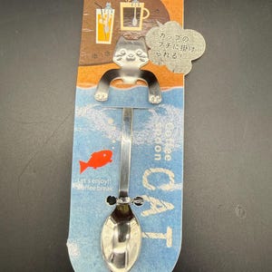 May include: A silver-toned metal spoon featuring a cat-shaped handle with extended arms designed to hang on a cup rim. The spoon is attached to a cardboard backing with blue and orange graphics, a red fish icon, and text including "Coffee spoon CAT" and "Let's enjoy!! Coffee break."