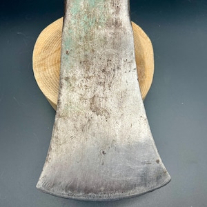 May include: A metal axe head with a weathered, silver-toned surface and green patina accents. The top portion features the stamped text Genuine Norlund. The metal shows signs of wear, including scratches and surface texture across the blade.