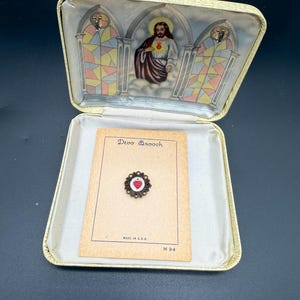 May include: An open, vintage, cream-colored jewelry box with a religious theme. Inside, a small brooch with a red heart design rests on a tan card. The box lid features a depiction of Jesus and stained-glass windows.