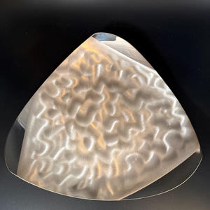 May include: A brushed metal, triangular serving tray with a wavy, textured surface. The tray has a smooth, curved edge and is set against a black background. The tray's design is modern and minimalist.