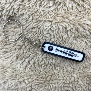 May include: A rectangular keychain with a black border and a white centre featuring a Spotify code. The keychain is attached to a silver chain and a large silver ring. The background is a textured, light brown fabric.