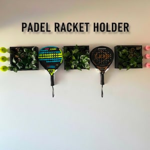 May include: A wall-mounted padel racket holder with the text "PADEL RACKET HOLDER". The holder features three black square planters with greenery, two padel rackets, and three yellow padel balls. The rackets have black handles.