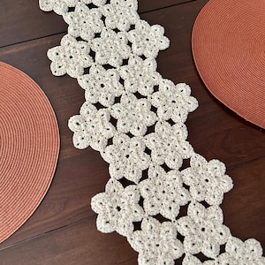 May include: A cream-colored, crocheted table runner with a floral pattern. The runner is placed on a dark brown wooden table, with two rust-colored placemats on either side. The runner's design features repeating flower shapes.