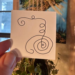 May include: A white square sticker with a black line drawing of a spiral and loops. The sticker is held up in front of a blurred background with a painting and greenery. The design is simple and minimalist.