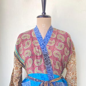 May include: A long-sleeved robe featuring a patchwork design with various floral and paisley patterns in shades of red, blue, green, and yellow. The garment includes a wrap-front closure with a matching fabric tie belt and two front pockets.
