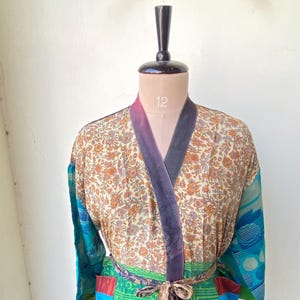 May include: A long-sleeved kimono-style robe featuring a cream-colored upper section with a dense orange and brown floral pattern, contrasted by a vibrant green lower section with geometric and floral motifs, a dark purple collar, and a tie belt.
