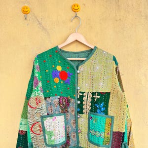 以下が含まれることがあります： 緑、オリーブ、タンの色合いのパッチワークジャケット。丸いネックラインと2つのフロントポケットが付いています。 ジャケットは、さまざまな模様の生地の正方形、装飾的なステッチ、ボタン留めが特徴です。 ジャケットは木製のハンガーに掛かっています。