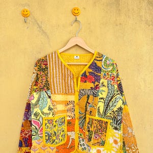 以下が含まれることがあります： 黄色、オレンジ、緑色の花柄、ストライプ、アニマルプリントを組み合わせたパッチワークジャケット。 ジャケットは丸襟、フロントポケット2つ、裏地は黄色です。 黄色の壁に木製のハンガーに掛けられています。