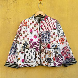 May include: A quilted jacket with a patchwork design featuring floral and geometric patterns in white, pink, purple, and blue. The jacket has a mandarin collar, two pockets, and black and white striped trim. It hangs on a wooden hanger.