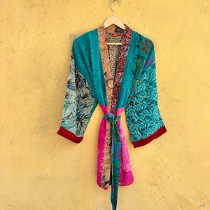 Può includere: Una vestaglia kimono in patchwork caratterizzata da un mix vivace di pannelli in tessuto color ottanio, magenta e rosso con vari motivi floreali e astratti. Il capo presenta maniche ampie, una cintura in vita coordinata e una fodera interna.