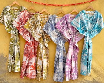 May include: A collection of colorful, patterned kaftans hanging against a yellow wall. The kaftans feature a palm tree design in various colors, including green, red, yellow, blue, and purple. Each kaftan is hanging on a wooden hanger.