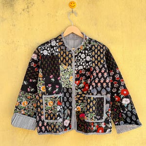 May include: A quilted patchwork jacket featuring floral patterns in red, white, orange, and green on a black base. The jacket has a collared neckline, two front pockets, and striped cuffs and trim. It is displayed on a wooden hanger.