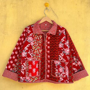 May include: A red quilted jacket with a floral pattern and striped accents. The jacket features a patchwork design with two front pockets and a collared neckline. The jacket is hanging on a wooden hanger against a yellow background.