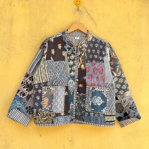 Peut inclure: Veste patchwork avec col officier, composée de carrés de tissu à motifs variés dans les tons de bleu, gris et marron. La veste est suspendue à un cintre en bois sur un mur jaune.