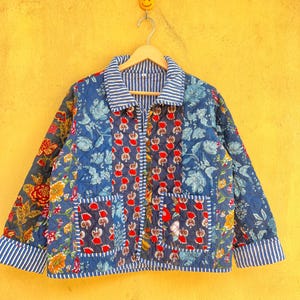 May include: A quilted jacket with a floral design in blue, red, and yellow. The jacket has a striped collar, cuffs, and pocket trim. It is suspended on a wooden hanger against a yellow backdrop.