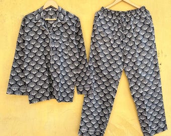 Op de afbeelding: Een marineblauwe pyjamaset met een wit waaiervormig patroon, bestaande uit een overhemd met knopen en een broek. Witte biezen omzomen de kraag, manchetten en broekzomen. De set hangt aan houten kleerhangers tegen een gele achtergrond.