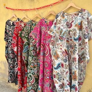 Puede incluir: Una fila de coloridos caftanes estampados colgados de perchas de madera. Los caftanes presentan diseños florales y de cachemira en varios colores, incluyendo negro, rojo, verde, rosa y blanco. El fondo es una pared amarilla.
