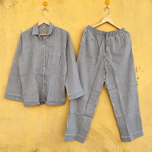 May include: A striped pyjama set in grey and white. The long-sleeved shirt has a chest pocket and buttons. The trousers have a drawstring waist. Both items have white trim and are displayed on wooden hangers.