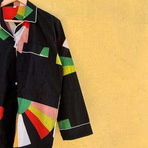 May include: Black pajama top with a colorful geometric design. The long-sleeved shirt features a white collar, cuffs, and pocket trim. The design includes red, green, yellow, pink, and white shapes. The shirt is hanging on a wooden hanger.