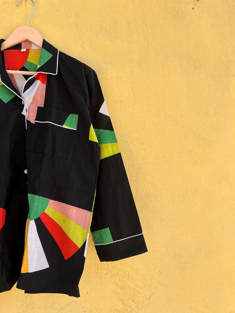 May include: Black pajama top with a colorful geometric design. The long-sleeved shirt features a white collar, cuffs, and pocket trim. The design includes red, green, yellow, pink, and white shapes. The shirt is hanging on a wooden hanger.