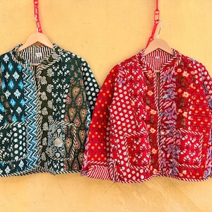 May include: Two patchwork quilted jackets hang on wooden hangers. One is dark green with blue and white patterns, the other is red with white polka dots and floral designs. Both have pockets and are displayed against a yellow wall.
