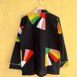May include: Black long-sleeved jacket with colorful geometric shapes in red, yellow, green, and white. The jacket has a white collar and trim. The garment is hanging on a metal hanger against a yellow wall.
