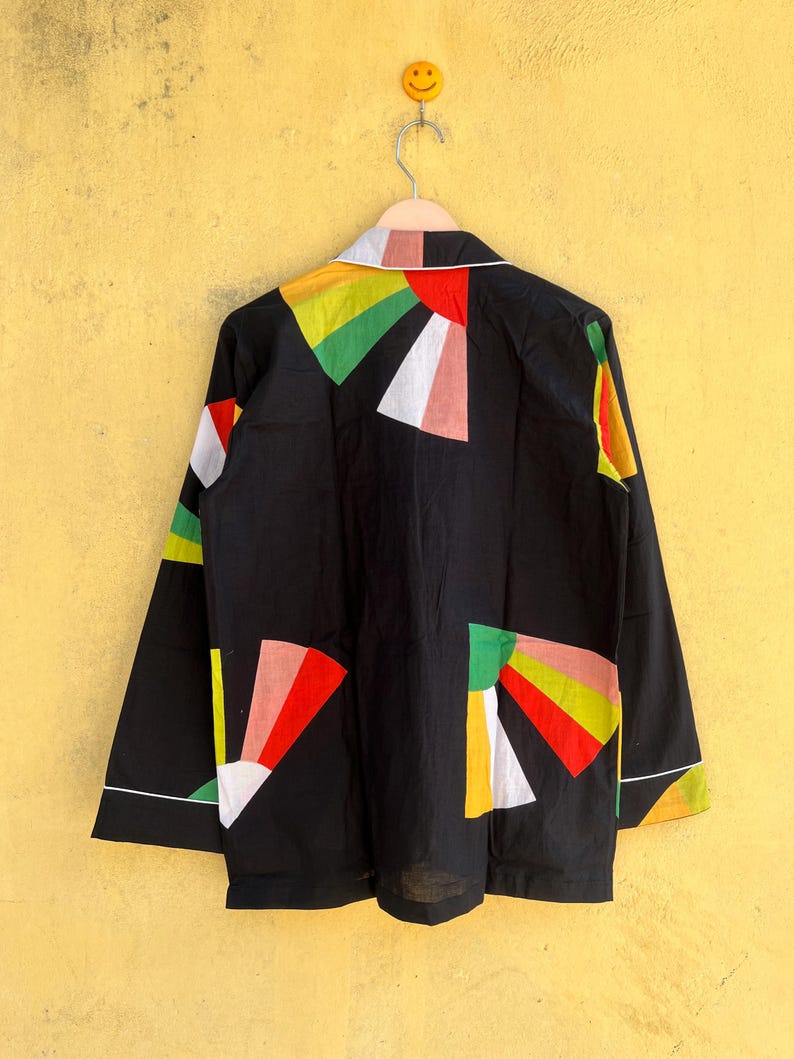 May include: Black long-sleeved jacket with colorful geometric shapes in red, yellow, green, and white. The jacket has a white collar and trim. The garment is hanging on a metal hanger against a yellow wall.