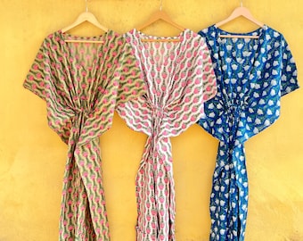 May include: Three patterned kaftans hanging against a yellow wall. The kaftans are in shades of pink, green, and blue, each with a unique floral design. They are displayed on wooden hangers, showcasing their flowing silhouettes.