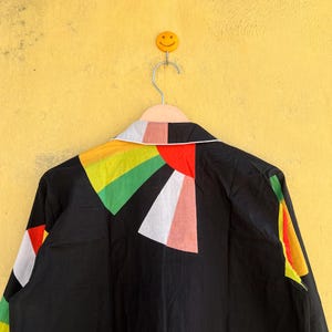 May include: Black jacket with a colorful geometric design on the back. The design features triangles in red, white, green, yellow, and pink. The jacket is hanging on a hanger against a yellow wall.