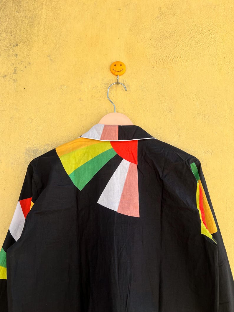 May include: Black jacket with a colorful geometric design on the back. The design features triangles in red, white, green, yellow, and pink. The jacket is hanging on a hanger against a yellow wall.