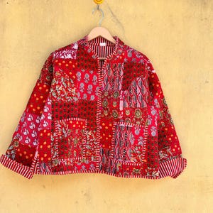 May include: A red patchwork jacket with various floral and geometric patterns. The jacket has a stand-up collar, two front pockets, and a striped lining. The cuffs are folded up, revealing a striped pattern. The jacket is hanging on a wooden hanger.