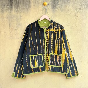 May include: A quilted jacket featuring a black base with vertical yellow and orange tie-dye stripes. The jacket has a mandarin collar, long sleeves, and two front patch pockets, all finished with lime green piping and a matching lime green interior lining.