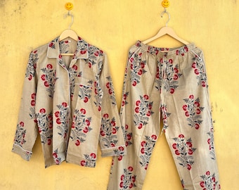 May include: A beige pajama set with a floral pattern of red flowers and dark green leaves. The long-sleeved top has a button-down front and a chest pocket. The pants have a drawstring waist and a relaxed fit. Both pieces have white trim.