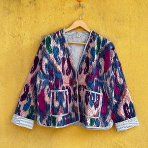 Puede incluir: Chaqueta acolchada con cuello en V, con un colorido estampado abstracto en tonos azules, rosas, verdes y morados. La chaqueta tiene dos bolsillos delanteros y está forrada con una tela a rayas. Cuelga de una percha de madera sobre un fondo amarillo.