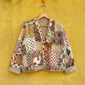May include: A patchwork jacket with a V-neck and button closure. The jacket features a variety of patterned fabric squares in shades of beige, red, blue, and white. The jacket is hanging on a wooden hanger against a yellow wall.