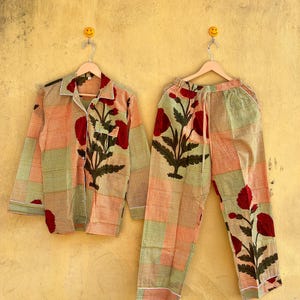 Peut inclure: Un ensemble de pyjama assorti, composé d'une chemise boutonnée et d'un pantalon. L'ensemble est orné d'un motif patchwork orange, vert et blanc, avec de grands motifs floraux rouges. Les pyjamas sont suspendus à des cintres en bois.