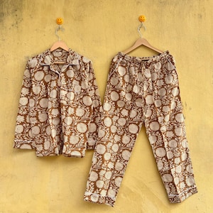 Peut inclure: Ensemble de pyjama à imprimé floral marron et crème. Le haut à manches longues a une fermeture boutonnée et un col. Le pantalon a une taille élastique. L'ensemble est suspendu à un cintre en bois sur fond jaune.