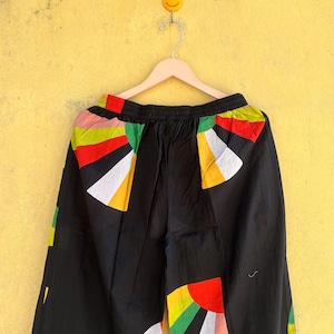 May include: Black pants with colorful geometric patterns in red, yellow, green, and white. The pants are hanging on a wooden hanger against a yellow wall. The design is a mix of abstract shapes.