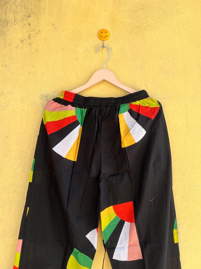 May include: Black pants with colorful geometric patterns in red, yellow, green, and white. The pants are hanging on a wooden hanger against a yellow wall. The design is a mix of abstract shapes.