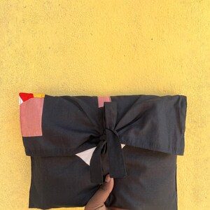 May include: A black fabric clutch with a bow closure. The clutch has a flap with geometric color block details in red, pink, and yellow. The background is a textured yellow wall.