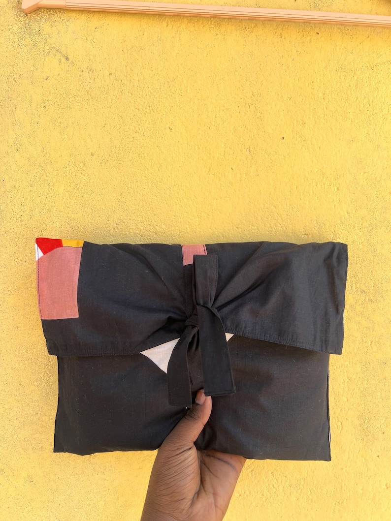 May include: A black fabric clutch with a bow closure. The clutch has a flap with geometric color block details in red, pink, and yellow. The background is a textured yellow wall.